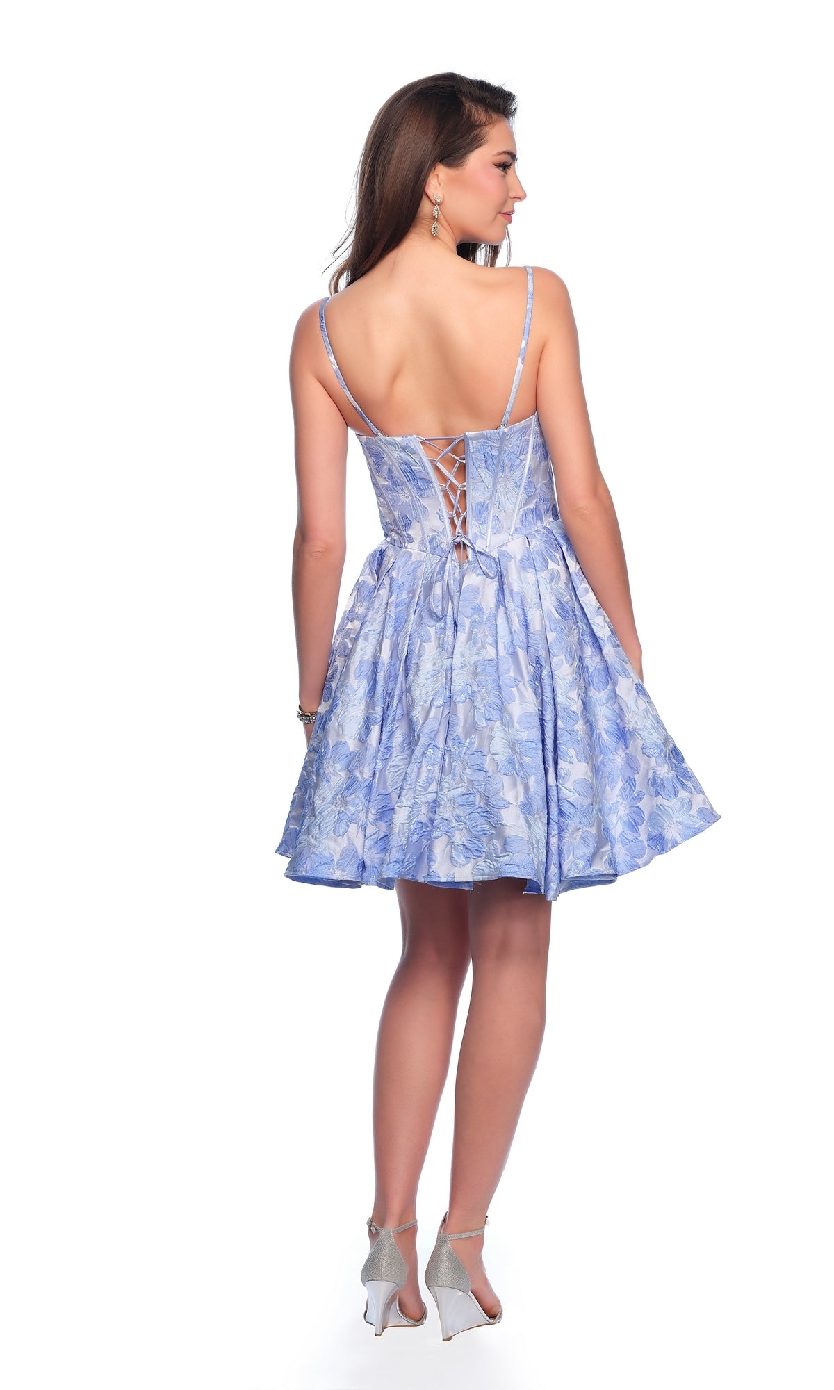A woman faces away from the camera in the Dave & Johnny Blue Floral Homecoming Dress 11960, featuring a lace-up corset back and paired with silver heels against a white background.
